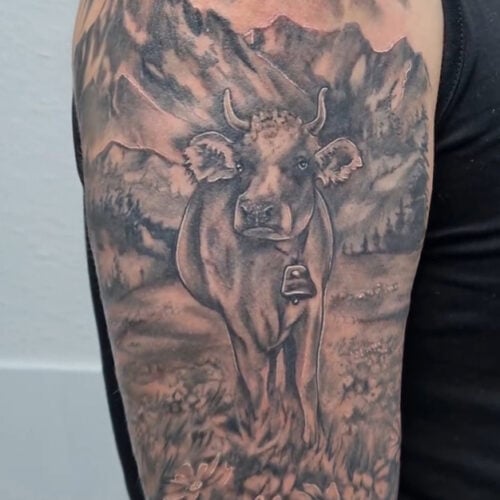 Tattoo oberarm mit weide bergpanorama und einer kuh im realistik stil